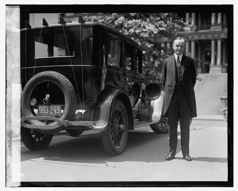 Calvin Coolidge stands beside an automobile fitted with a roof antenna.