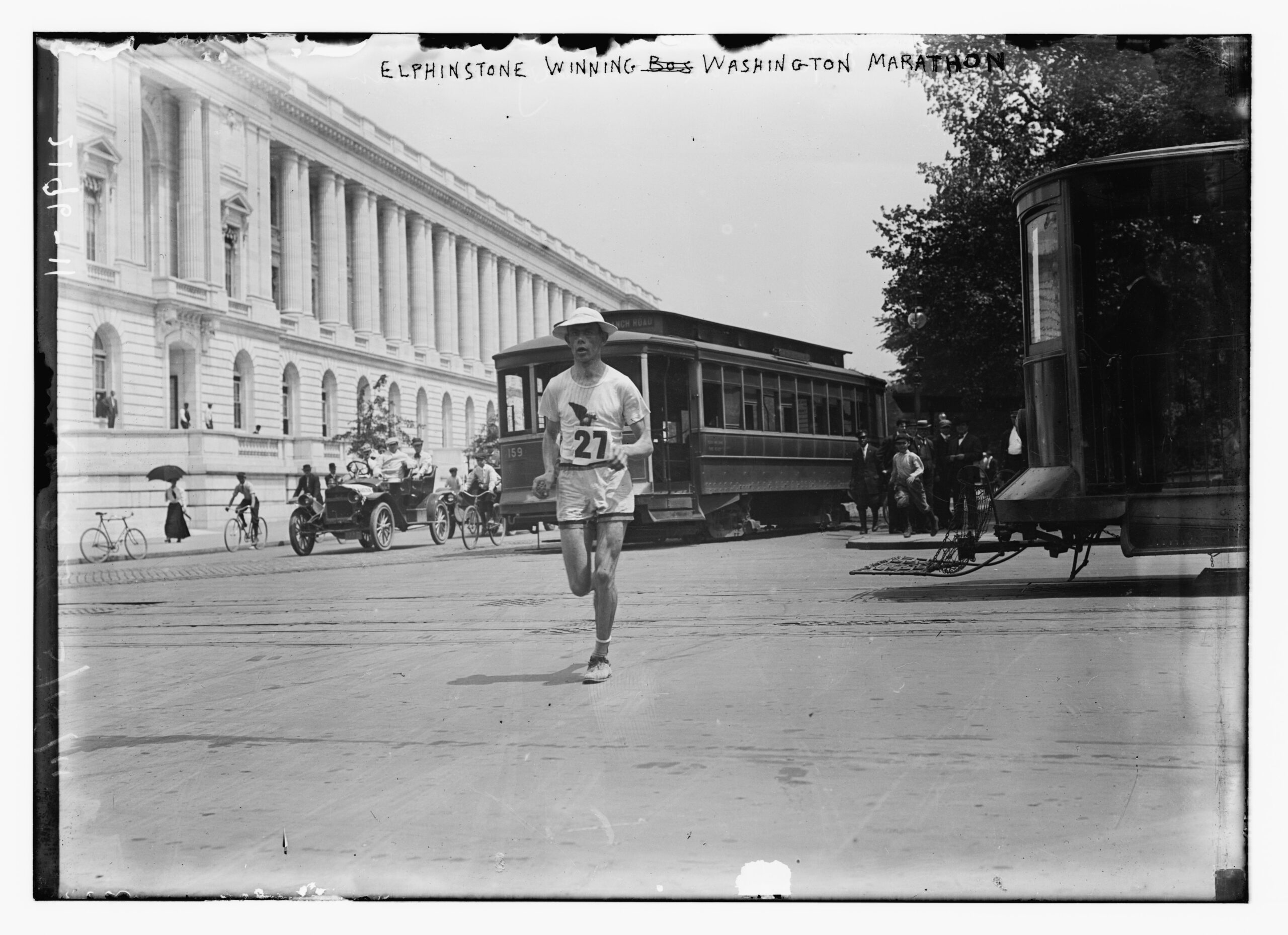 Henry Elphinstone, wearing race number 27, runs past streetcars and spectators.