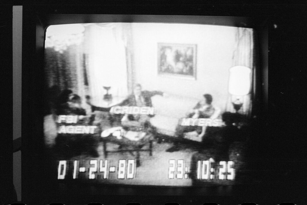 A television screen showing grainy black-and-white FBI surveillance footage of Rep. Michael Myers and others seated in a room during the Abscam sting, 1980.