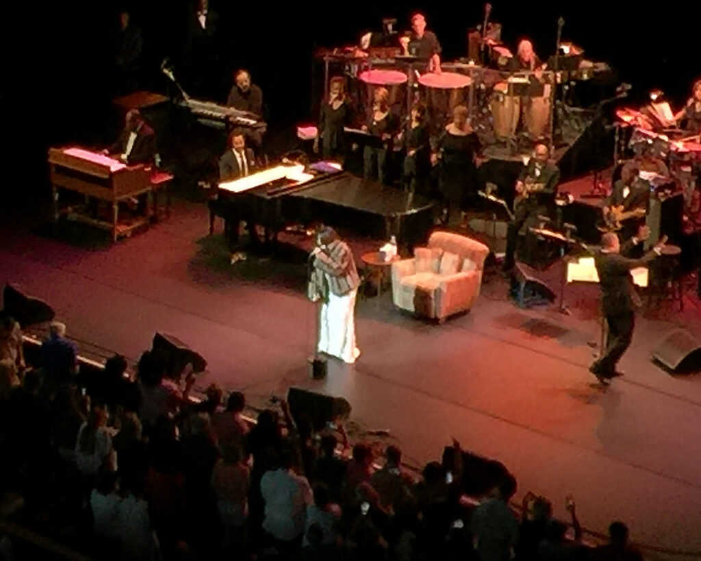 Aretha Franklin in a white gown at the front of the Filene Center stage, with her band behind her.