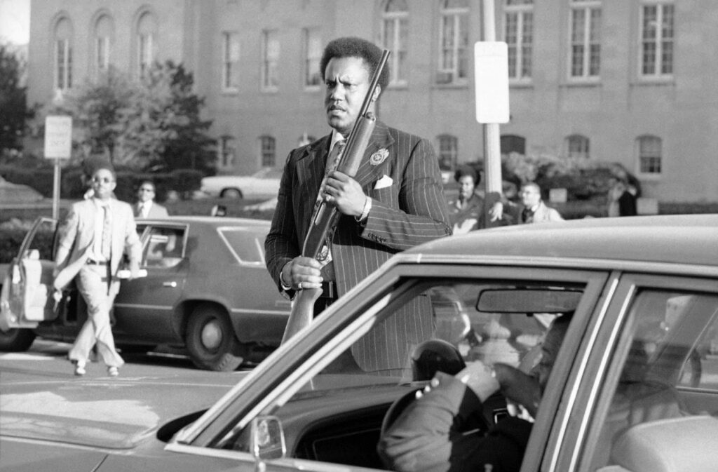 A man in a pinstripe suit with a badge on his lapel holding a shotgun upright beside a car in traffic.