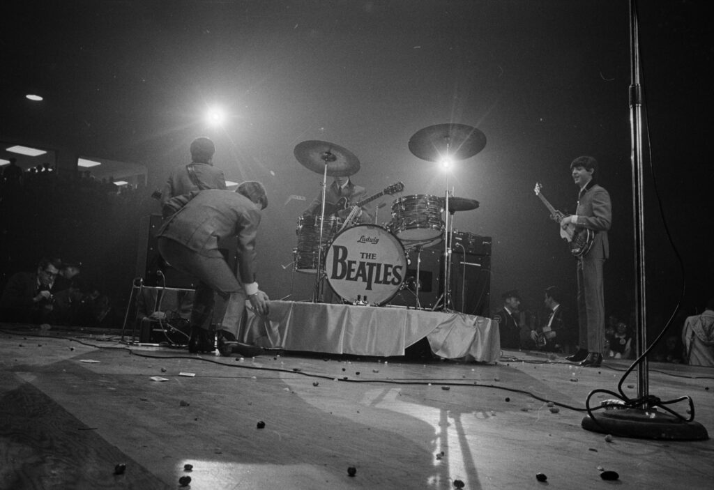 The Beatles on the center stage under spotlights, a man bent over the drum riser with The Beatles drum head, small debris scattered on the stage floor