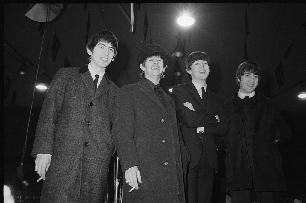 The four Beatles in overcoats and suits, smiling under arena lights, two of them holding cigarettes