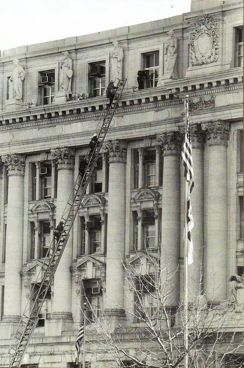 Firefighters climbing a long extension ladder against the columned stone facade of the District Building on Pennsylvania Avenue.
