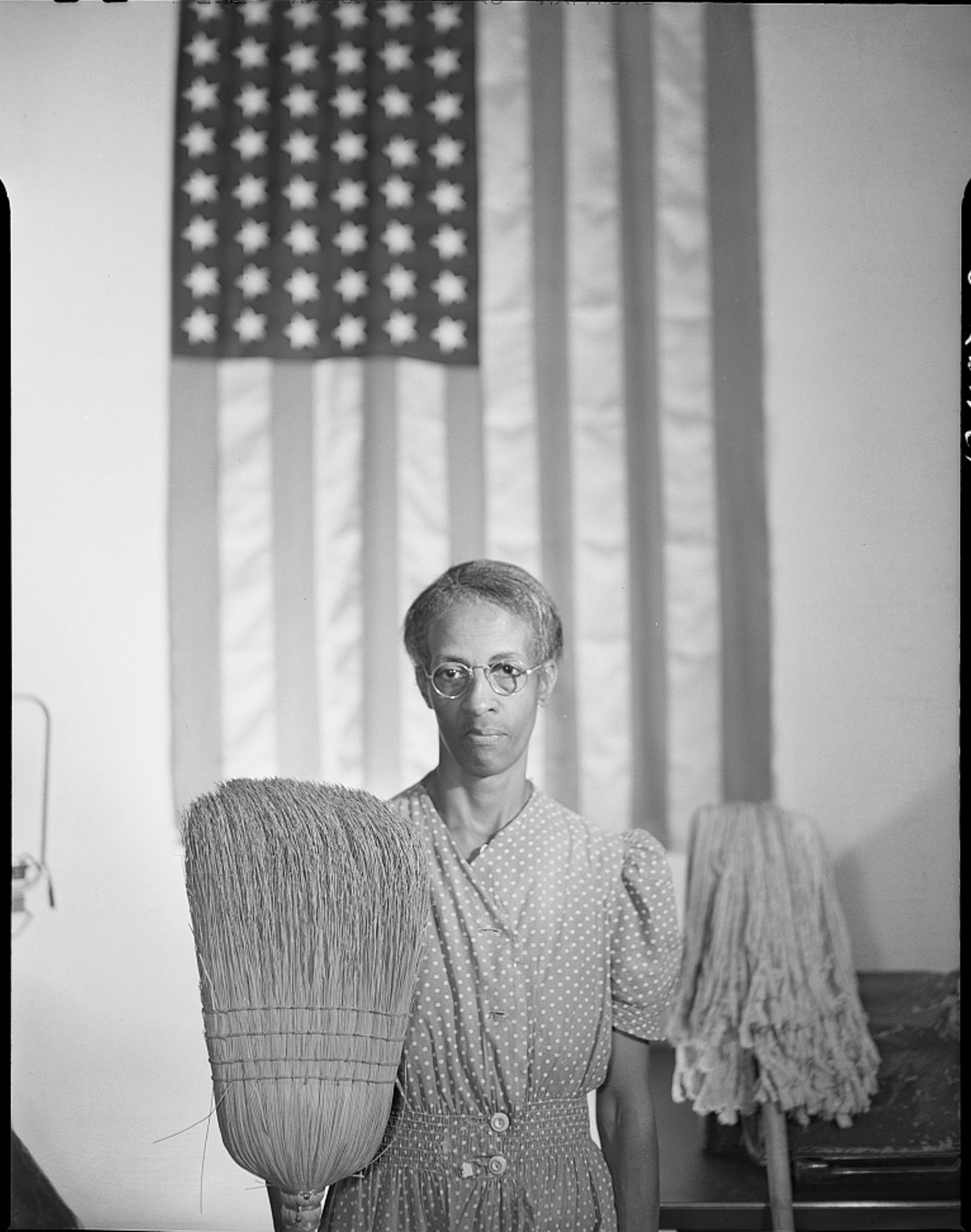 Ella Watson stands before an American flag holding a broom and mop, Gordon Parks, 1942