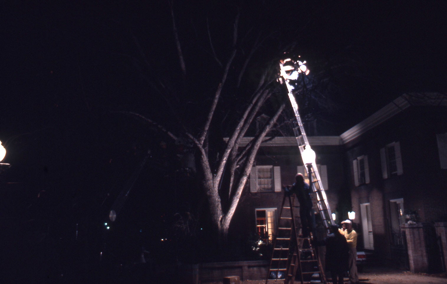 A film crew on ladders beside a lit streetlamp in front of a brick house on Prospect Street at night
