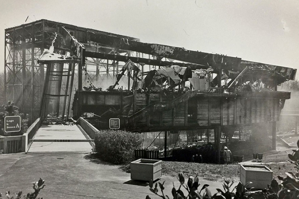 The burned-out frame of the original Filene Center, its roof collapsed and smoke still rising.