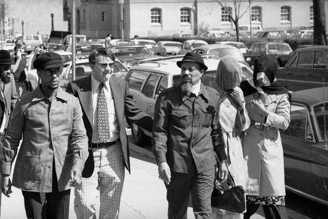 Hamaas Abdul Khaalis, bearded and wearing a hat, walking across a parking lot with several men and two women in head scarves.