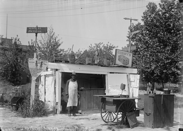 A man in a long white apron stands in front of a small wooden lunch stand covered with hand-painted signs