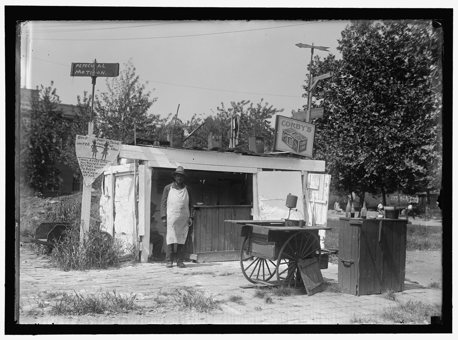 A man in a long white apron stands in the doorway of a small wooden lunch stand covered with hand-painted signs