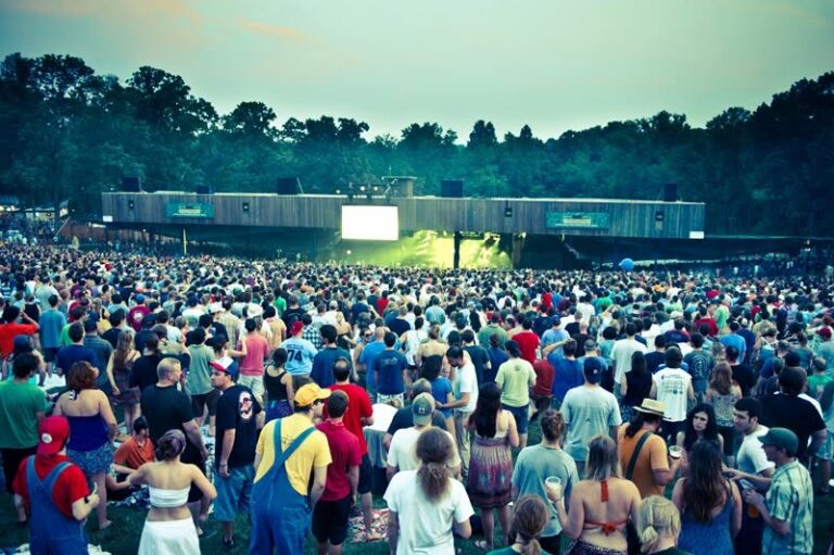 A packed lawn of concertgoers at dusk facing the lit Merriweather Post Pavilion stage, with trees behind.