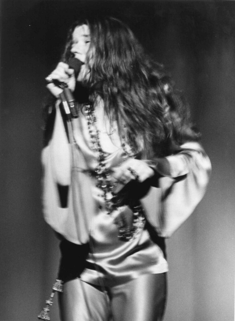 Janis Joplin sings into a microphone onstage, eyes closed, wearing beads and a satin top.