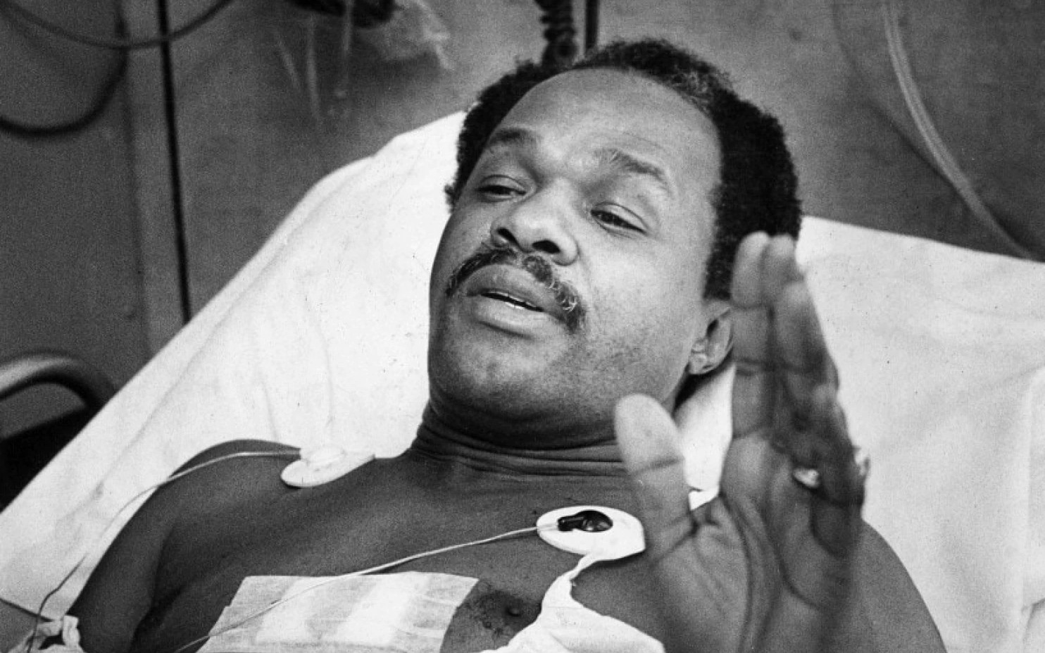 Councilman Marion Barry lying in a hospital bed with a bandage and monitor leads on his chest, gesturing as he speaks.