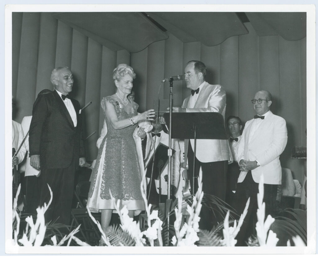 Marjorie Merriweather Post in a formal gown and Vice President Hubert Humphrey exchange a ticket onstage, with Jim Rouse and conductor Howard Mitchell standing nearby.
