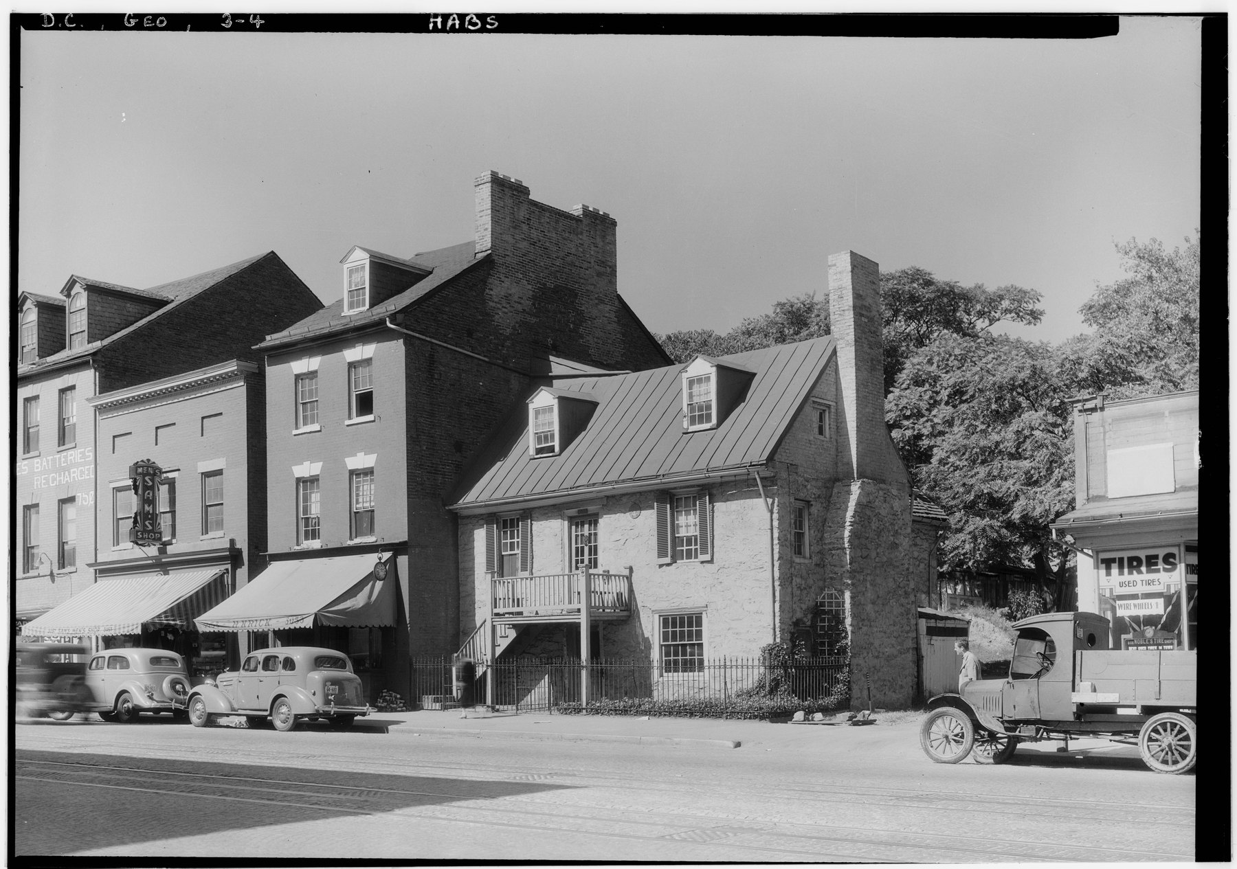 1935 street view of M Street in Georgetown with the Old Stone House set between brick storefronts, parked cars and a tire shop sign
