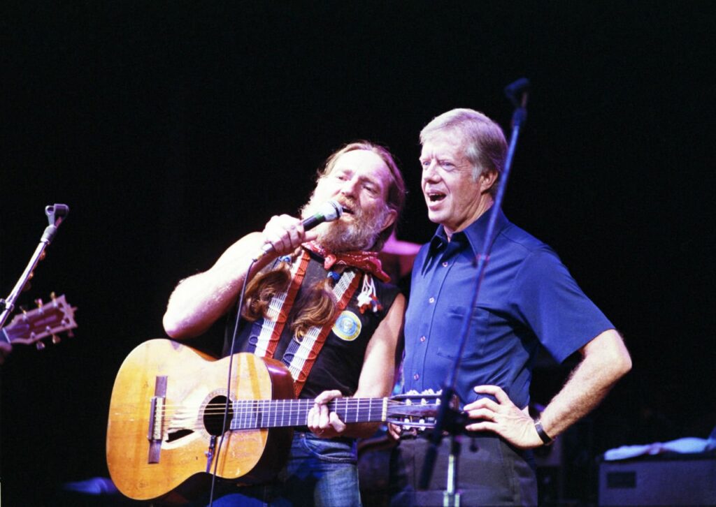 Willie Nelson sings into a microphone with his guitar while President Jimmy Carter stands beside him onstage.