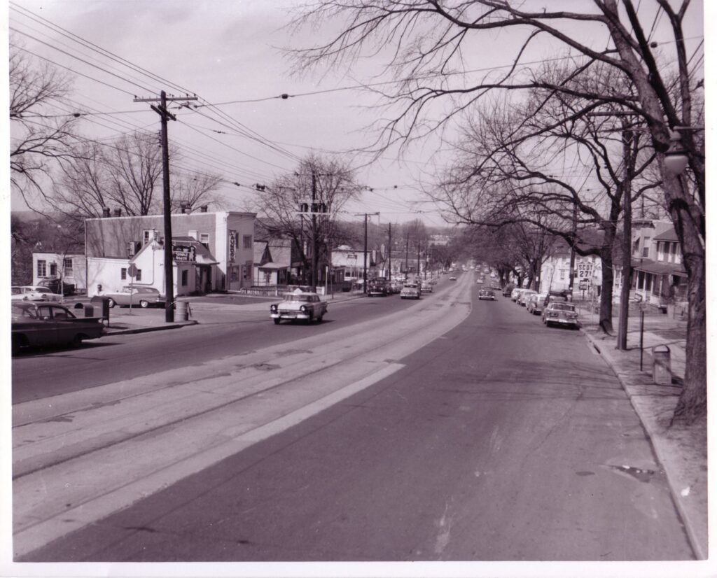 Wisconsin Avenue NW at Chesapeake Street around 1960, with streetcar tracks in the center lane, overhead trolley wires, a Toddle House and a gas station sign reading 27 cents