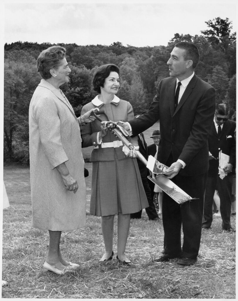 Catherine Filene Shouse, Lady Bird Johnson and Stewart Udall stand in a field holding a ceremonial shovel.