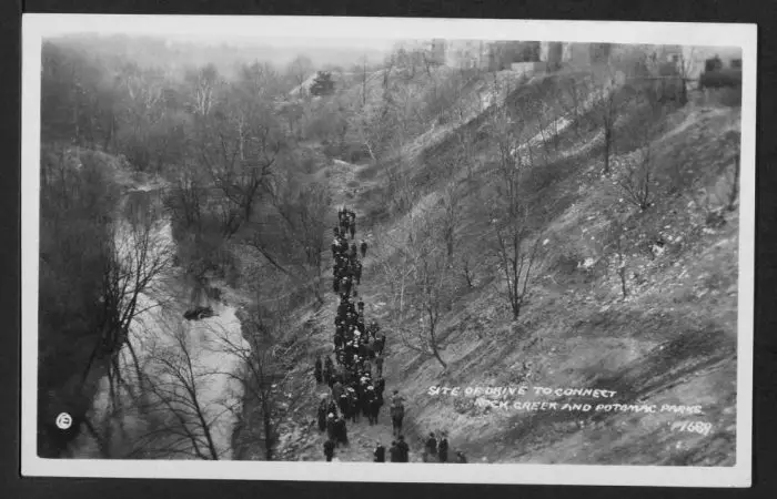 View of a group of hikers walking alongside Rock Creek to the left and an embankment that leads up to the right upon which can be seen a row of buildings. Location is to the west and downhill from the 2800 block of Adams Mill Road NW.